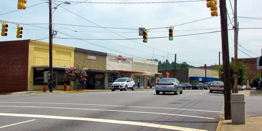 Main Street in downtown Taylorsville, North Carolina: a row of low brick and painted storefronts, a crape myrtle in pink bloom, traffic lights over the street and wooded hills behind